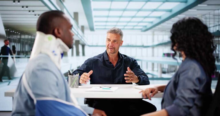 A person reviews medical bills and financial documents at a kitchen table with a supportive friend nearby.